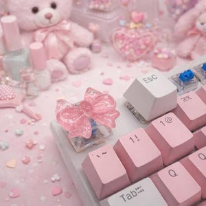 May include: A pink bow-shaped keycap sits on a white keyboard with pink keys. The keycap is translucent and has a button in the center. Other items in the background include nail polish, a teddy bear, and heart-shaped decorations.