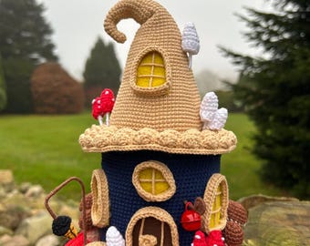 May include: A whimsical, crocheted fairy house with a tan pointed roof and a navy blue base. The house features yellow-framed windows, a brown door, and is adorned with crocheted mushrooms and other decorative elements. It sits on a red base.