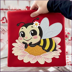 May include: A red square textile featuring an embroidered cartoon bee resting on a daisy. The bee has black and yellow stripes, large eyes, and a flower on its head. The daisy has pink and white petals and a yellow center.