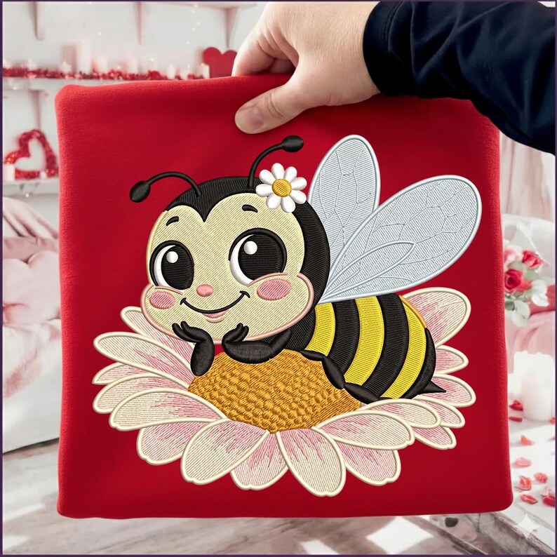 May include: A red square textile featuring an embroidered cartoon bee resting on a daisy. The bee has black and yellow stripes, large eyes, and a flower on its head. The daisy has pink and white petals and a yellow center.