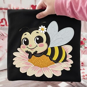 May include: Black square fabric featuring an embroidered cartoon bee with a daisy on its head, resting on a pink and yellow flower. The bee has black and yellow stripes and large, friendly eyes. The fabric is held by a person.