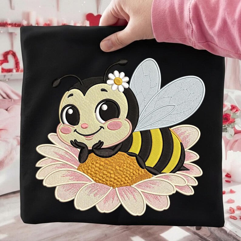May include: Black square fabric featuring an embroidered cartoon bee with a daisy on its head, resting on a pink and yellow flower. The bee has black and yellow stripes and large, friendly eyes. The fabric is held by a person.