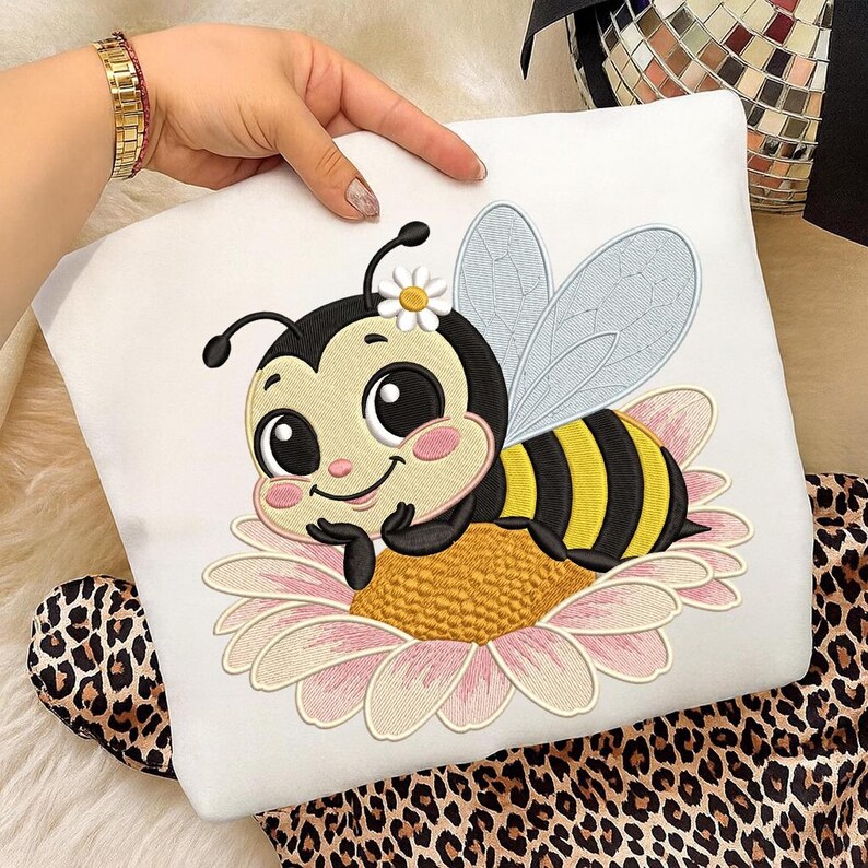 May include: White pillow with an embroidered cartoon bee design. The bee has a black and yellow striped body, light blue wings, and a daisy on its head. The bee is resting on a pink and yellow flower.