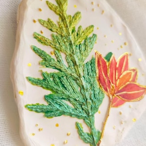 Könnte beinhalten: Handgefertigter, cremefarbener Ornament mit strukturierter Oberfläche. Es zeigt ein detailliertes, grün und gold bemaltes Blatt und eine rot-orangefarbene Blume. Goldglitzer akzentuiert das Design, und eine goldene Schnur ist zum Aufhängen befestigt.