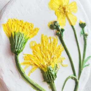 Op de afbeelding: Een wit, ovaal ornament met een reliëf van gele bloemen en groene stelen. De bloemen zijn gedetailleerd en geplaatst tegen een getextureerde achtergrond. Een gouden koord is bevestigd om op te hangen.