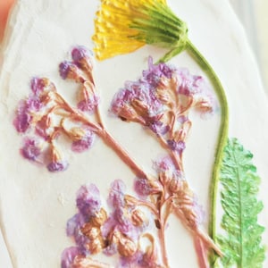 Op de afbeelding: Een witte, rechthoekige, handgemaakte gipsen ornament met geperste bloemen. Het ontwerp omvat een gele paardenbloem, paarse bloemen en groene bladeren. Een gouden koord is bevestigd om op te hangen.