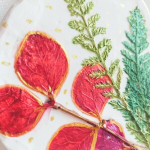 Op de afbeelding: Een wit keramisch ornament met een botanisch ontwerp. Voorzien van rode bladeren met gouden rand, groene varen- en groenblijvende takjes en gouden spikkels. Een gouden koord is bevestigd om op te hangen.