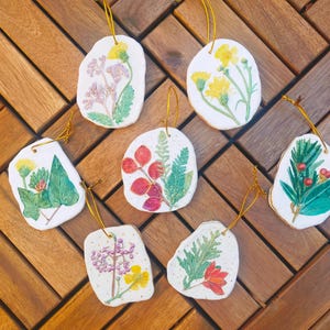 May include: Seven handmade ceramic ornaments in irregular shapes. Each features a unique watercolour botanical design, including flowers and foliage, with gold string for hanging. The ornaments are arranged on a wooden surface.