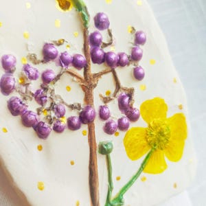 Op de afbeelding: Handgemaakte, crèmekleurige keramische ornament met een bloemenontwerp. Het ornament heeft een bruine stengel met paarse bessen, een gele bloem en gouden accenten. Een gouden koord is bevestigd om op te hangen.