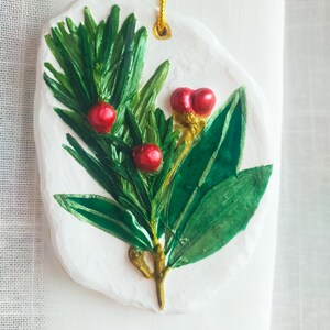 Op de afbeelding: Een handgemaakte kerstversiering met een geschilderd botanisch ontwerp. De versiering heeft een witte basis met een groen en rood plantenontwerp. De plant heeft groene bladeren en rode bessen, met gouden accenten en een gouden ophangkoord.