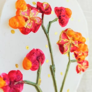 May include: A white, teardrop-shaped ceramic ornament featuring a floral design. The flowers are red and orange with gold accents, and the background has gold polka dots. A natural twine loop is at the top.