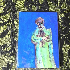 May include: A blue postcard featuring a pastel drawing of a ghostly figure in a green coat. The figure has pale skin and crossed arms. The postcard is in a clear plastic sleeve.