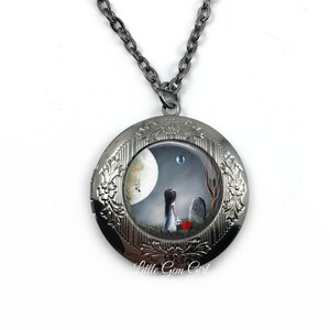 Könnte beinhalten: Eine silberne Halskette mit einem Anhänger in Form eines Medaillons, auf dem ein gemaltes Bild einer Frau vor einem Grabstein unter einem Halbmond zu sehen ist. Die Frau hält ein rotes Herz in der Hand. Der Anhänger ist mit einem floralen Muster am Rand verziert.