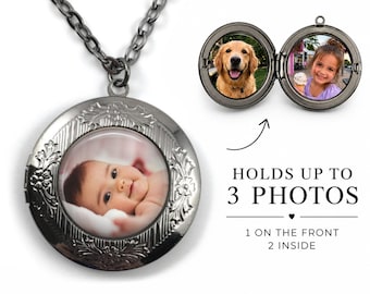 May include: A round gunmetal locket necklace with an ornate floral border. The front displays a baby photo, while the interior reveals two additional circular photo frames containing a dog and a child. Text reads: HOLDS UP TO 3 PHOTOS 1 ON THE FRONT 2 INSIDE.