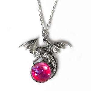 May include: Silver dragon pendant necklace featuring a red and purple orb. The dragon has detailed wings and hangs from a delicate chain. The pendant is a fantasy-themed accessory, suitable for everyday wear.