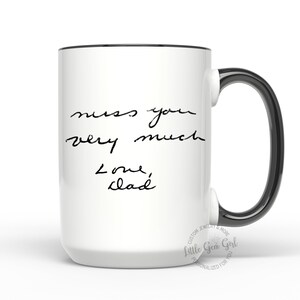 Może przedstawiać: Biały ceramiczny kubek z czarną obw&oacute;dką i uchwytem. Kubek zawiera odręczny tekst, kt&oacute;ry brzmi "miss you very much Love, Dad". Kubek to spersonalizowany prezent.