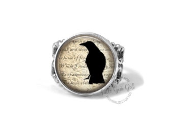 Op de afbeelding: Een zilveren ring met een zwart raafsilhouet op een vintage papierachtergrond. De ring heeft een ronde glazen cabochon met een zwart raafsilhouet op een vintage papierachtergrond. De tekst op het papier luidt "upon a dmy weak and wear volume of for While I nodo As of someone Tis some visi and in".
