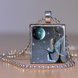 Könnte beinhalten: Eine silberne Kettenkette mit einem quadratischen Anhänger, der ein Gemälde einer blauen Fee zeigt, die auf einem Ast sitzt, mit einem großen blauen Mond im Hintergrund.