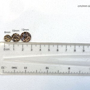 May include: Three round, flat, decorative embellishments in different sizes, 8mm, 10mm, and 12mm, with a black and gold speckled pattern, are lined up on a clear ruler with centimeter and millimeter markings.