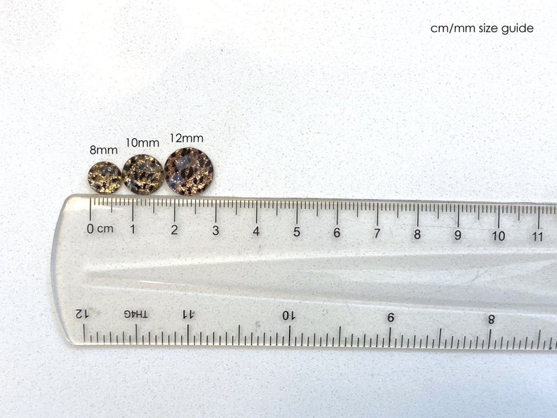 May include: Three round, flat, decorative embellishments in different sizes, 8mm, 10mm, and 12mm, with a black and gold speckled pattern, are lined up on a clear ruler with centimeter and millimeter markings.