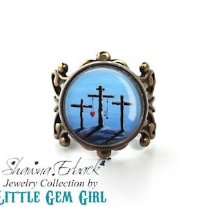 Könnte beinhalten: Ein Ring mit einem runden, gläsernen Cabochon, der ein Gemälde von drei Kreuzen vor blauem Himmel zeigt. Die Kreuze sind braun und der Himmel ist hellblau. Ein kleines rotes Herz ist an der Basis der Kreuze gemalt. Der Ring ist aus Messing und hat ein dekoratives Design.