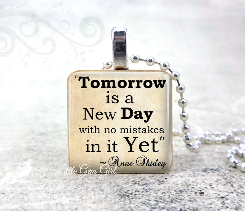 May include: A square pendant necklace featuring the quote "Tomorrow is a New Day with no mistakes in it Yet" by Anne Shirley. The pendant has a silver bail and hangs on a silver ball chain. The background is a soft, blurred white.