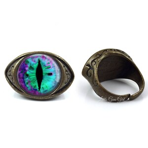 May include: A ring with a green, purple, and black eye design set in an oval-shaped, antique brass setting.