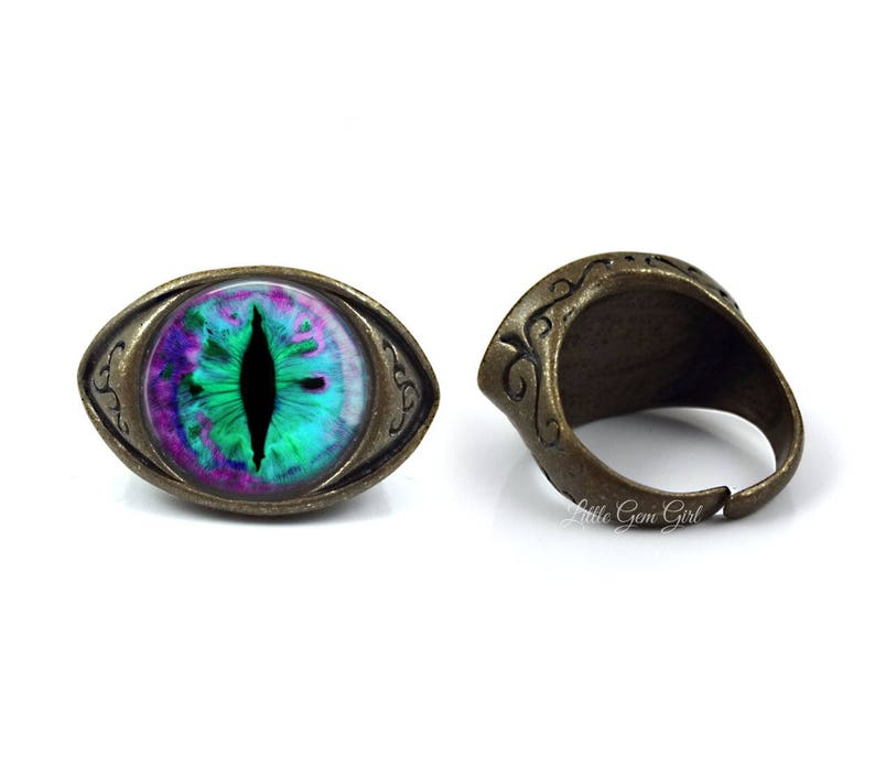 May include: A ring with a green, purple, and black eye design set in an oval-shaped, antique brass setting.