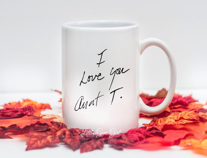 Może przedstawiać: Biały ceramiczny kubek z odręcznym napisem "I love you Aunt T." w kolorze czarnym. Kubek ma zakrzywiony uchwyt i stoi wśr&oacute;d czerwonych i pomarańczowych jesiennych liści. Słowa "Little Gem Girl" znajdują się na dole.