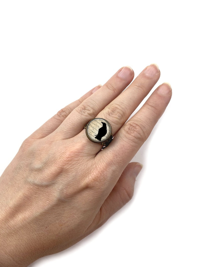 Pu&ograve; includere: Un anello in ottone con una silhouette di corvo nero su sfondo bianco. Il corvo &egrave; appollaiato su un ramo con un design di scrittura vintage.