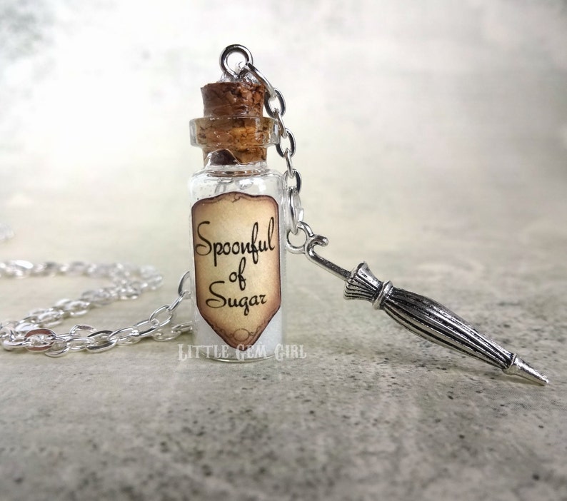 May include: A silver chain necklace with a small glass bottle charm. The bottle has a cork stopper and a label that reads "Spoonful of Sugar". The necklace also has a small silver charm shaped like an umbrella.