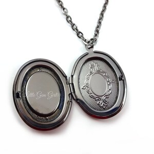 May include: A black metal locket necklace with an oval-shaped pendant. The locket is open, revealing a blank space inside. The locket has a decorative design on the back.