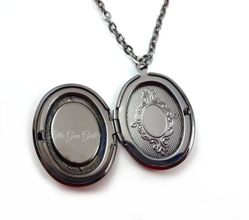 May include: A black metal locket necklace with an oval-shaped pendant. The locket is open, revealing a blank space inside. The locket has a decorative design on the back.