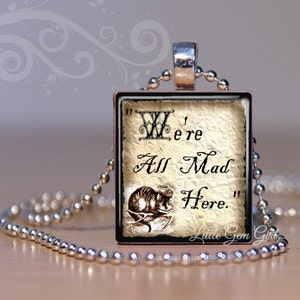 May include: A silver chain necklace with a square pendant featuring a vintage illustration of the Cheshire Cat from Alice in Wonderland. The pendant has a white background with black text that reads "We're All Mad Here."