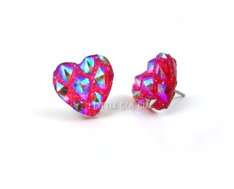 May include: A pair of heart-shaped earrings with a red base and iridescent glitter. The earrings are made of a clear material with a textured surface.