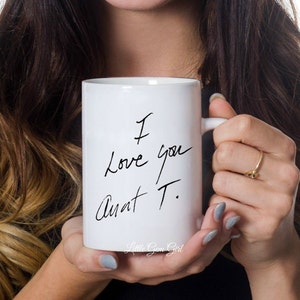 Może przedstawiać: Biały ceramiczny kubek z odręcznym napisem "I Love you Aunt T." czarnym pismem. Kubek ma uchwyt i jest trzymany przez osobę. Słowa "Little Gem Girl" znajdują się na dole kubka.