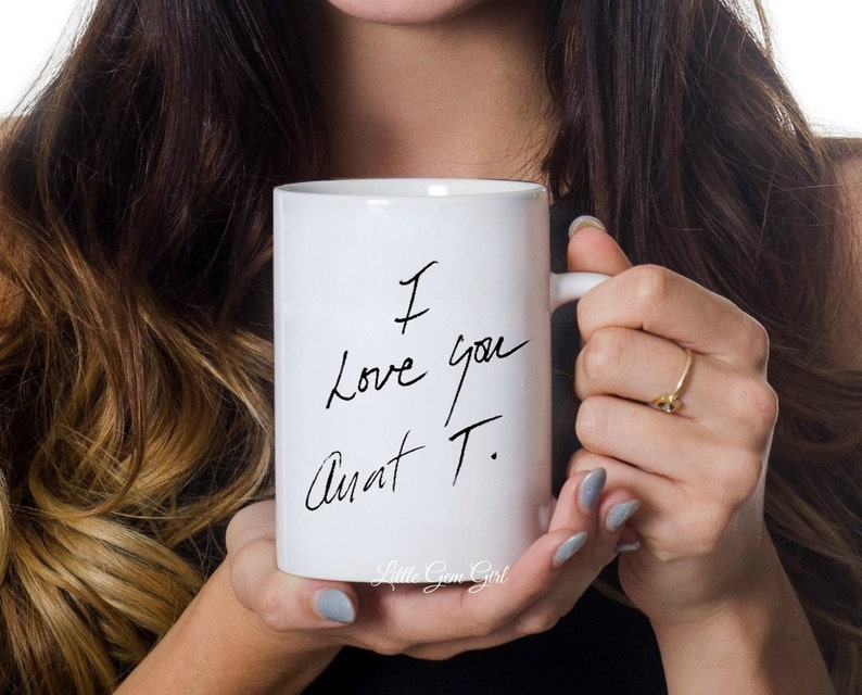 Może przedstawiać: Biały ceramiczny kubek z odręcznym napisem "I Love you Aunt T." czarnym pismem. Kubek ma uchwyt i jest trzymany przez osobę. Słowa "Little Gem Girl" znajdują się na dole kubka.