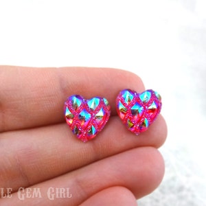 May include: A pair of pink heart-shaped earrings with a glittery, iridescent finish. The earrings have a geometric pattern and are a vibrant shade of pink.