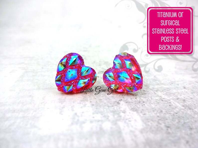 May include: A pair of heart-shaped earrings with a pink base and iridescent blue and green glitter. The earrings are made with titanium or surgical stainless steel posts and backings.