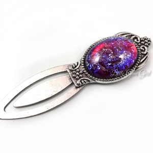 Könnte beinhalten: Silbernes Lesezeichen mit einem großen, ovalen, irisierenden Glascabochon. Der Cabochon ist eine Mischung aus roten, violetten und blauen Farben. Das Lesezeichen hat ein dekoratives, verziertes Design.