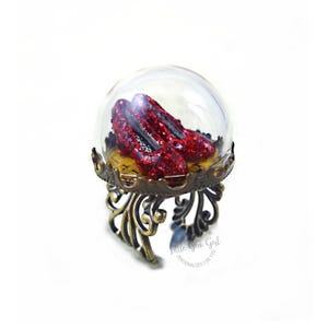 May include: A glass dome ring with a pair of red glitter shoes inside. The dome sits on a gold metal stand with a decorative design. The ring is a unique and whimsical piece of jewelry.