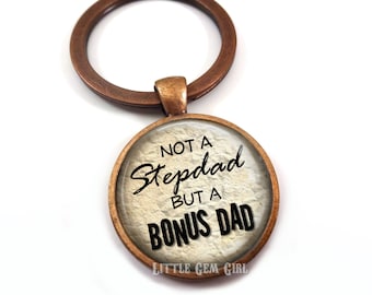 May include: A bronze-colored key chain with a round glass charm. The charm has a white background with black text that reads "Not a Stepdad But a Bonus Dad" and the text "Little Gem Girl" at the bottom.