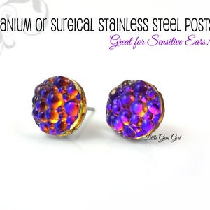 May include: Two stud earrings with a textured, iridescent surface in shades of purple, orange, and yellow. The earrings have a post backing and are set in a gold-colored metal. Text on the image reads "Titanium or Surgical Stainless Steel Posts" and "Great for Sensitive Ears!"
