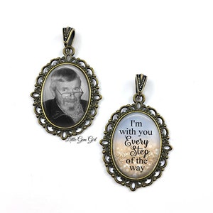 May include: Two bronze oval photo pendants with a decorative filigree border. One pendant features a black and white photo of a man with a beard and glasses. The other pendant has a blue and white background with gold glitter and the text "I'm with you every step of the way".