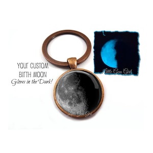 May include: A bronze-colored key chain with a round glass charm featuring a black and white image of the moon. The moon glows in the dark. The charm has the text "Little Gem Girl" on the bottom. The text "Your Custom Birth Moon Glows in the Dark!" is written below the key chain.