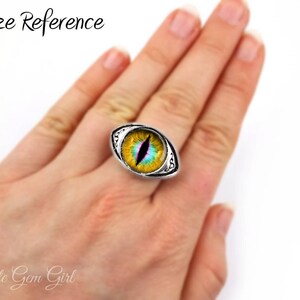 May include: A silver ring with a yellow, green, and blue eye design. The ring is on a person's finger, showing the size of the ring.