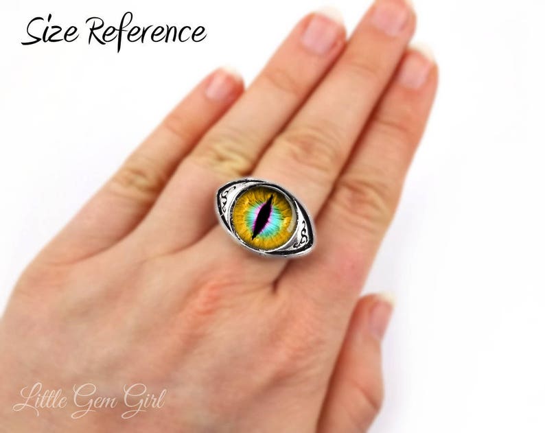 May include: A silver ring with a yellow, green, and blue eye design. The ring is on a person's finger, showing the size of the ring.