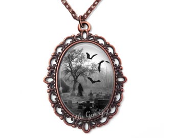 May include: A copper-toned necklace with an oval pendant featuring a black and white image of a graveyard with a lone figure walking through it. Bats fly overhead. The pendant has a decorative filigree frame and the words "Little Gem Girl" are printed on the bottom.