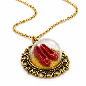 May include: A gold-toned necklace featuring a round pendant with an ornate filigree border. The pendant contains a clear glass dome enclosing a pair of miniature red glittery shoes resting on a yellow brick pattern background.