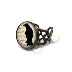 Pu&ograve; includere: Un anello regolabile in bronzo con un cammeo rotondo in vetro che presenta una silhouette nera di un corvo su uno sfondo di carta vintage. Il testo sulla carta recita "ak and wear", "lume of for", "While I nodo", "s of someone", "some visi".